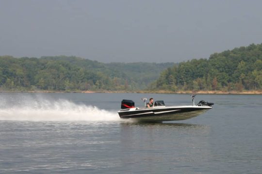 Bass boat with trolling motor speeding accross a lake with trees in the background.