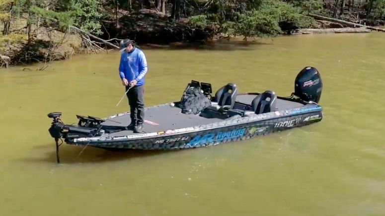 Bass fisherman looking at his fish finder and fishing at the front of his bass boat on muddy water, with woods in the background.