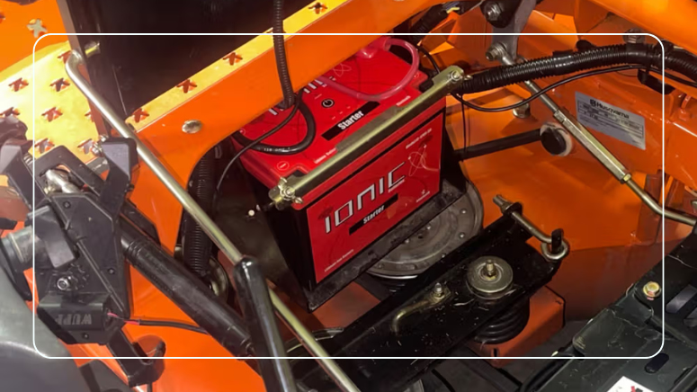 Red Ionic lithium starter battery installed in a riding mower battery compartment with cables and hold-down bracket