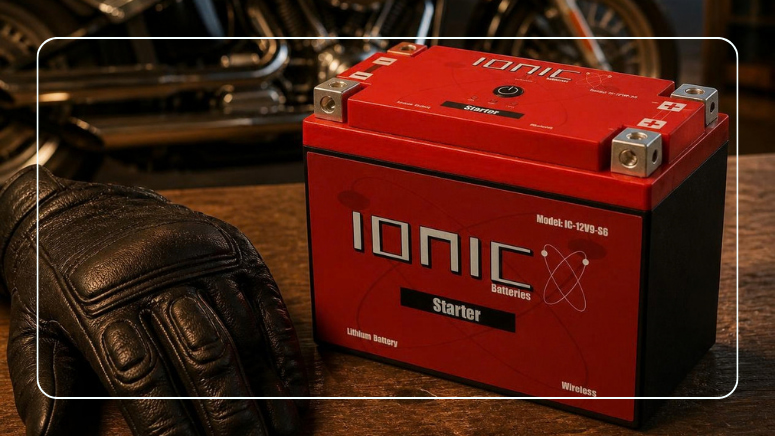 Ionic LiFePO4 motorcycle starter battery on a workbench next to leather riding gloves with a motorcycle in the background