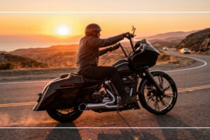 Rider on a black Harley-style touring motorcycle cruising a coastal highway at sunset