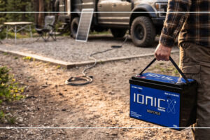 Person carrying Ionic Deep Cycle lithium battery at off-grid campsite with solar panel and 4x4 vehicle in background